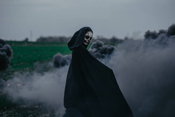 若い女性は黒煙を背景に死のハロウィーンの衣装のフィールドに立っています。ハロウィンのコンセプト。砂糖の頭蓋骨のメイク. - 写真・画像