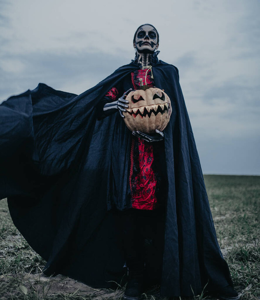 Mujer joven se encuentra entre el campo en el disfraz de Halloween de la muerte con esqueleto pintado en su cuerpo y jack-o-linterna contra el fondo cielo oscuro. Concepto de Halloween. Maquillaje de cráneo de azúcar. - Foto, imagen