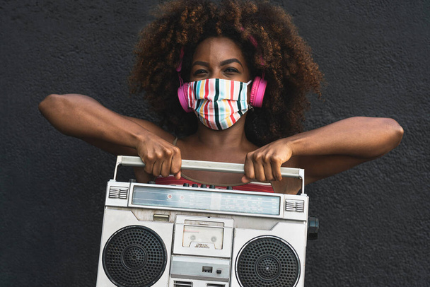 Молода африканка, одягнена в маску обличчя, танцює надворі, слухаючи музику за допомогою бездротових навушників і стерео бумбоксу. - Фото, зображення