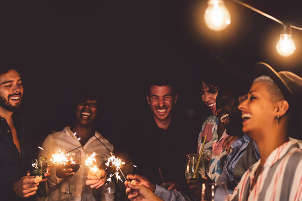 Gelukkig jonge vrienden vieren oudejaarsavond met sterretjes vuurwerk en cocktails drinken op patio house party - Jeugd mensen levensstijl en vakantie concept - Foto, afbeelding