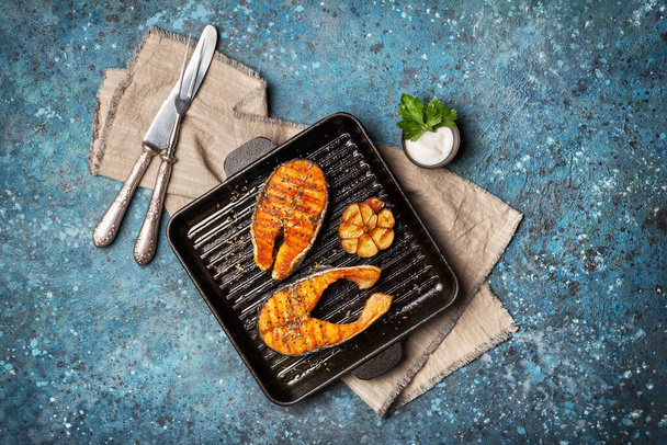 Top view of grilled red fish salmon steak on cast iron skillet or pan with spice, fried garlic and tasty sauce on blue concrete background - Foto, afbeelding
