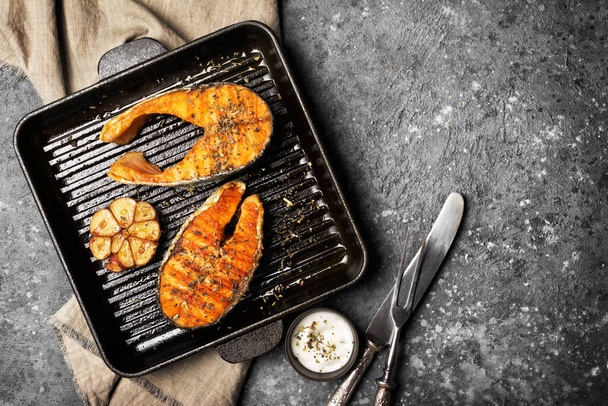 Top view of grilled red fish salmon steak on cast iron skillet or pan with spice, fried garlic and tasty sauce on grey concrete background - Foto, Imagem