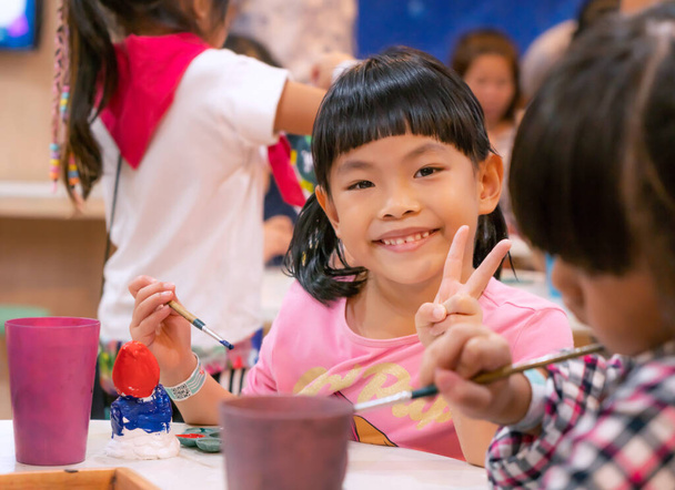 Portrét asijské dítě dívka v umělecké dílně, šťastný a usmívající se tvář, show gesto dva prsty, roztomilý obličej s černými vlasy a bang. Rozmazané popředí a pozadí spolužáka. - Fotografie, Obrázek