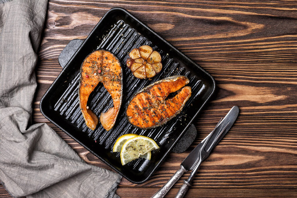 Top view of grilled red fish salmon steak on cast iron skillet or pan with spice, fried garlic and lemon on brown wooden background - Zdjęcie, obraz