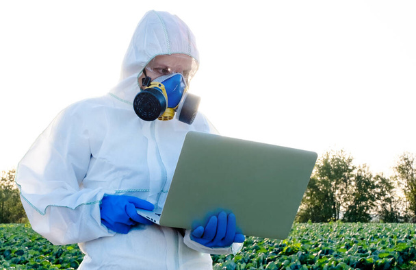Επιστήμονας φορώντας ένα λευκό προστατευτικό εξοπλισμό, χημική μάσκα και γυαλιά χρησιμοποιεί Laptop στο αγρόκτημα τομέα. - Φωτογραφία, εικόνα