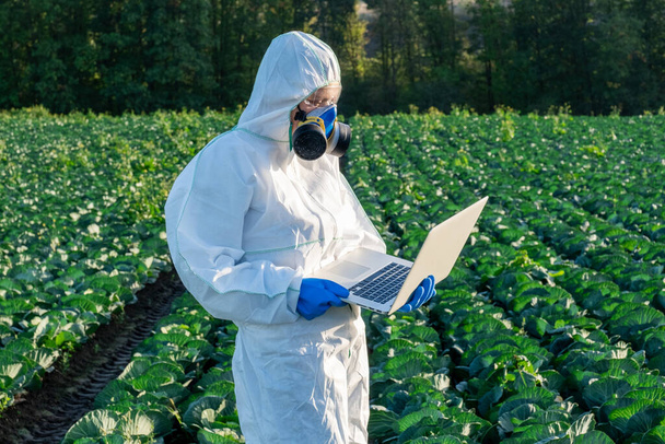 Fehér védőfelszerelést, kémiai maszkot és szemüveget viselő tudós laptopot használ a mezőgazdasági területen. - Fotó, kép