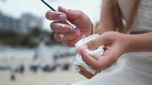Jovem loira pinta um quadro na praia da cidade. No ar livre Limpa a pintura de mãos - Filmagem, Vídeo