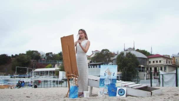 Joven rubia pinta cuadro en la playa Al aire libre En el fondo de las gaviotas voladoras - Imágenes, Vídeo