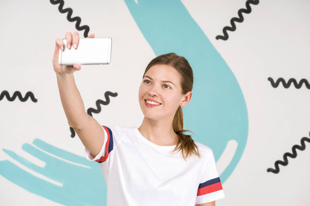 Νεαρή όμορφη γυναίκα στέκεται σε λευκό φόντο τέχνη με smartphone στο χέρι, λαμβάνοντας selfie φωτογραφία - Φωτογραφία, εικόνα