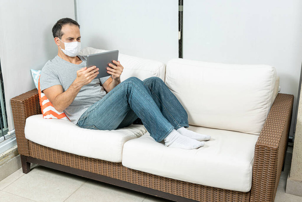 44-letni mężczyzna z maską oglądający tablet na kanapie. - Zdjęcie, obraz