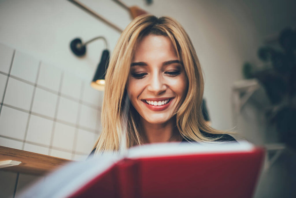 Happy Caucasian female student enjoying leisure time for reading literature book with positive plot, cheerful hipster girl holding education textbook and smiling while checking noted information - Photo, Image
