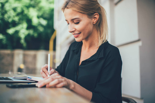 Happy female student with cute smile on face making information notes while learning at outdoors table desktop,cheerful Caucasian woman 20 years old writing data while accounting in education textbook - Valokuva, kuva