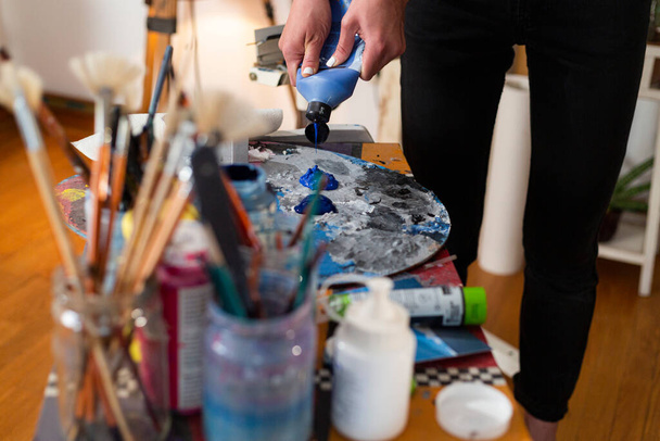 Close up of painter's hand extruding color on a palette in her studio. Around the palette are a multitude of brushes for painting in jars. - Photo, Image