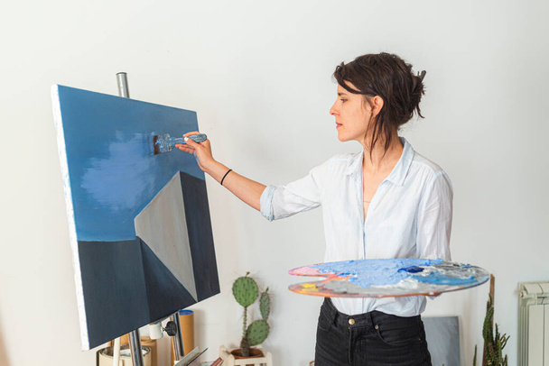 A young smiling painter applies paint to a canvas in her home studio. Other accompanying materials for painting could be seen around it. She holds a palette of paints in one hand, while with the other she applies the paint with a paintbrush on the ca - Photo, Image