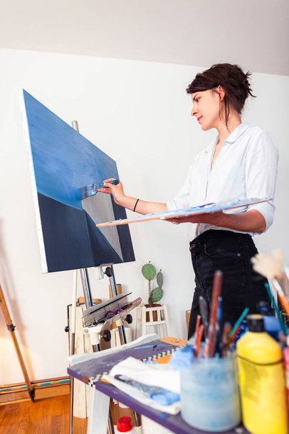 Μια νεαρή ζωγράφος που χαμογελάει βάζει μπογιά σε έναν καμβά στο στούντιο του σπιτιού της. Άλλα συνοδευτικά υλικά για τη ζωγραφική θα μπορούσε να δει γύρω από αυτό. Κρατάει μια παλέτα χρωμάτων στο ένα χέρι, ενώ με το άλλο απλώνει το χρώμα με ένα πινέλο - Φωτογραφία, εικόνα