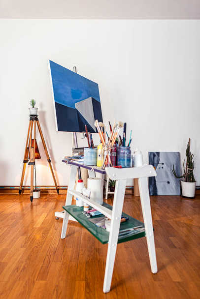 A painters work of art in studio. Around you can see a contemporary work of art placed on an easel, other pieces of art, as well as other accompanying material for painting. - Photo, Image