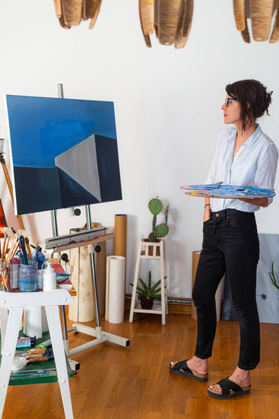 A young painter observes her work of art in her home studio. Around her, you can see a contemporary work of art placed on an easel, other pieces of art, as well as other accompanying material for painting. - 写真・画像