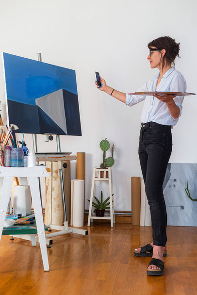Una joven pintora con teléfono móvil en el estudio de su casa. Fotografia su obra de arte con un teléfono móvil, mientras sostiene una paleta de colores en su otra mano. - Foto, Imagen