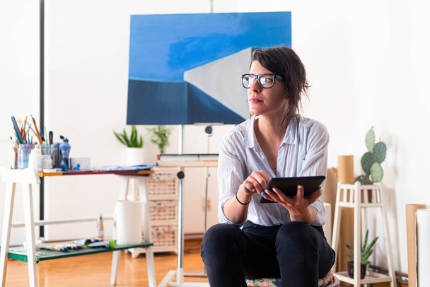 Mladá malířka sedí ve svém domácím studiu a používá tablet jako inspiraci. Nosí brýle, zatímco doprovodný materiál pro malování je vidět kolem ní v pozadí. - Fotografie, Obrázek