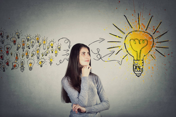 Thoughtful student girl, keeps hand under chin, looking up positive thinking for a solution. Ingenious young woman teenager gathering new ideas into a big lightbulb. Genius creativity concept. - Photo, Image