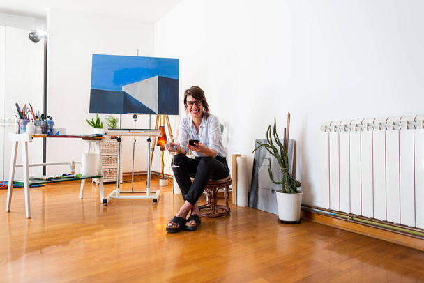 Una joven pintora está sentada en el estudio de su casa leyendo algo de su teléfono móvil. Ella está usando gafas y sosteniendo una copa de vino blanco, mientras que el material de acompañamiento para la pintura se puede ver a su alrededor en el fondo. - Foto, imagen