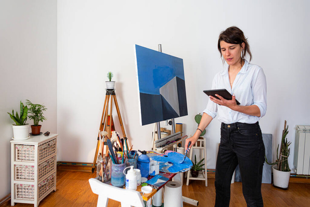 The young painter is standing in her home studio and copying her work from her tablet directly on the canvas. She is holding a tablet in one hand and a paintbrush in the other. Around it in the background you can see the accompanying material for pai - Photo, Image