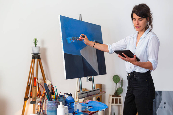 The young painter stands in her home studio and copies the sketch from her tablet directly on the canvas.Around it in the background you can see the accompanying material for painting. - Photo, image