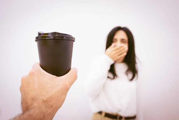 De cerca la mano sostiene la taza de café negro para llevar que ofrece la mujer empleada bostezando en fondo blanco de la pared. - Foto, Imagen