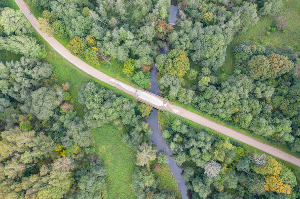 Εναέρια πάνω προς τα κάτω θέα του χωματόδρομου και της γέφυρας πάνω από τον ποταμό περιέλιξης που ρέει μέσα από πράσινο δάσος - Φωτογραφία, εικόνα