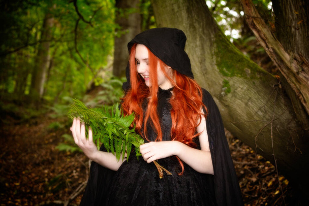 Gyönyörű fiatal nő hosszú vörös hajjal, fekete köpenyben az erdőben, kezében zöld páfránnyal. - Fotó, kép