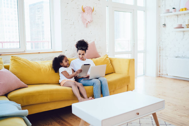 African American kid showing tablet to interested mom in jeans and white shirt while sitting on yellow couch at home together - 写真・画像