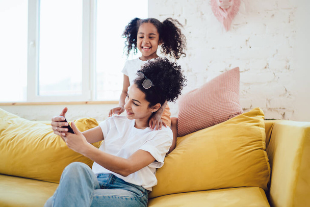 Black young cheerful woman taking selfie with laughing funny kid with pigtails behind back on yellow home couch with pillows - Foto, afbeelding