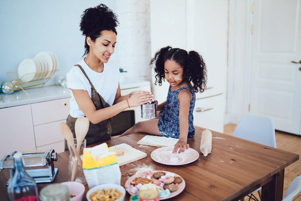 African American brunette daughter sitting on table and taking last pinch of flour near mother in white shirt and apron in kitchen - Foto, afbeelding