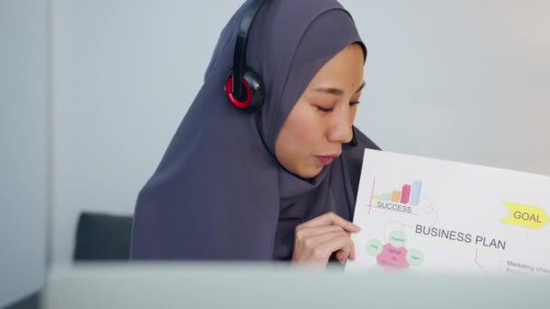 Азиатская мусульманка носит наушники, используя ноутбук, чтобы поговорить с коллегами о продаже отчета в конференц-видео-звонке во время работы из домашнего офиса по ночам. Социальное дистанцирование, карантин для коронного вируса. - Кадры, видео