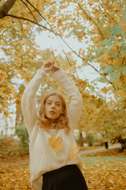 Nő az őszi parkban. Egy tinédzser lány az őszi levelek hátterében. Divatfotó egy fehér meleg pulóveres modellről. Szőke a kamerába néz. - Fotó, kép