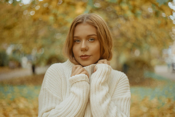 Photo portrait of a girl in a white sweater, the blonde holds the neck of the sweater and looks at the camera. Pretty woman in the park in autumn. Autumn fashion and style - Photo, Image