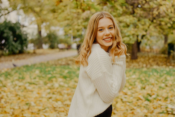 Ένας έφηβος τρέχει στο πάρκο και γελάει, το φθινόπωρο έχει έρθει. Φθινοπωρινά φύλλα και δέντρα σε πράσινο φόντο. Το κορίτσι είναι χαρούμενο, χαμογελαστό και γλυκό - Φωτογραφία, εικόνα