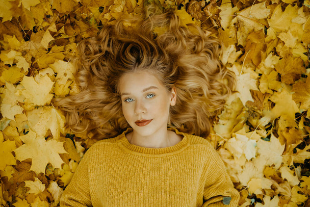 美しい少女は秋に公園にいます。オレンジの葉を背景に黄色いセーターを着た10代。ブロンドはカメラを見ている。ファッションとスタイル - 写真・画像