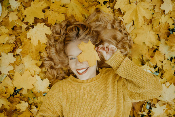 Beautiful girl lies in the park in autumn. A teenager in a yellow sweater on a background of orange leaves. The blonde smiles and laughs. Fashion and Style. Woman covering eyes with leaf and smiling - Photo, Image