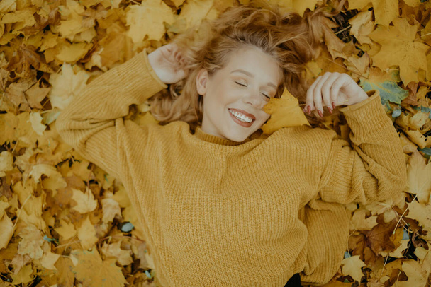 Beautiful girl lies in the park in autumn. A teenager in a yellow sweater on a background of orange leaves. The blonde smiles and laughs. Fashion and Style - Photo, Image