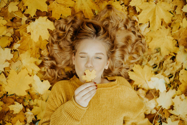 Beautiful girl lies in the park in autumn. A teenager in a yellow sweater on a background of orange leaves. The blonde smiles and laughs. Fashion and Style. Woman blowing a kiss - Photo, Image