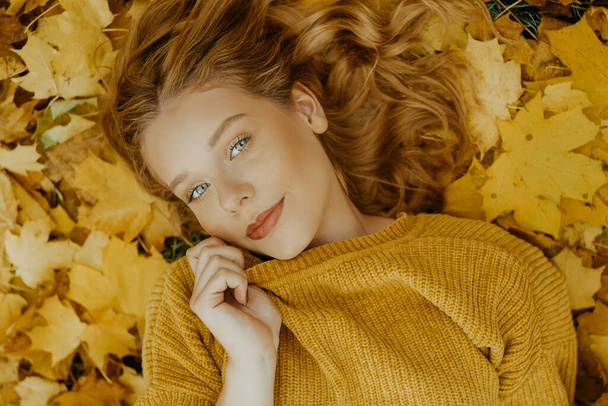 Gyönyörű lány fekszik a parkban ősszel. Egy sárga pulóveres tinédzser narancssárga levelek hátterében. A szőke a kamerába néz. Divat és stílus - Fotó, kép