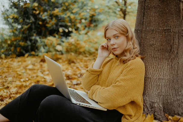 A beautiful girl teenager sits in an autumn park with a laptop. The student studies on the street. Study in the fall. The woman looks into the camera. - Valokuva, kuva