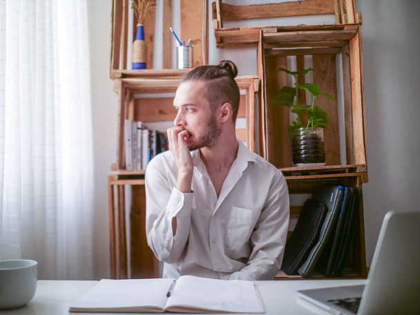 jonge blanke man in wit shirt zitten aan een bureau is zenuwachtig bijten zijn nagels - Foto, afbeelding