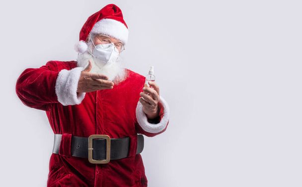 Père Noël portant un masque, pointant bouteille de gel hydroalcoolique en fond blanc - Photo, image