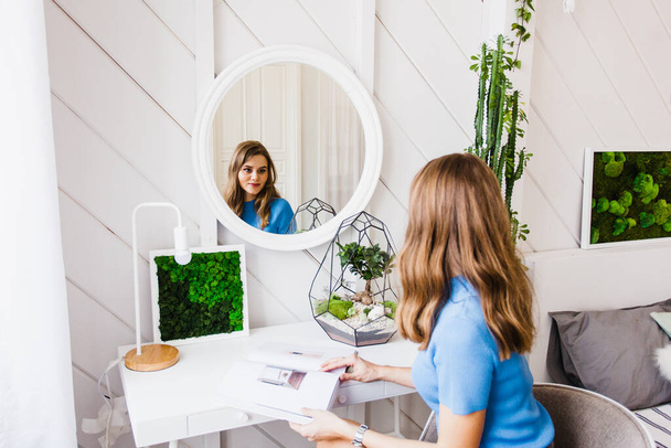 The girl is sitting near the mirror, leafing through a magazine, a beautiful bright interior with a bed, a picture of moss, plants - Фото, изображение