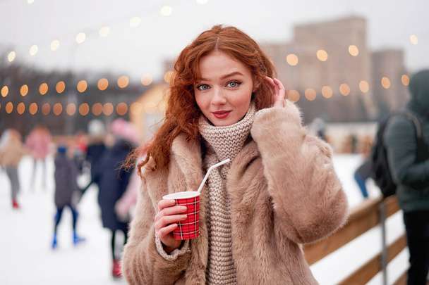 Young beautiful redhead girl freckles ice rink background Pretty woman curly hair portrait walking new year fair Modern cute female drink hot beverage mulled wine chrismas holiday decorated street.  - Photo, image
