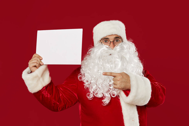 Surprised Santa Claus points to a blank white plate for text. Red studio background - Fotoğraf, Görsel