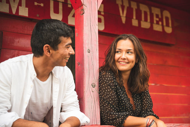 Bonita pareja coqueteando y sonriendo unos a otros fuera de un salón de madera roja. Fotografía del concepto de rancho - Foto, imagen