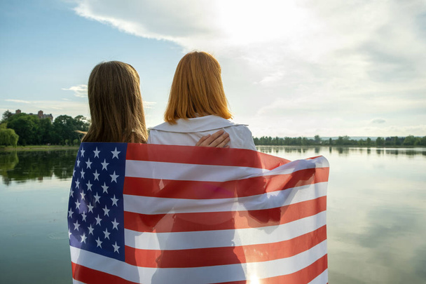 Dwie młode przyjaciółki z flagą USA na ramionach przytulające się do siebie na świeżym powietrzu nad brzegiem jeziora. Patriotyczne dziewczyny świętują dzień niepodległości Stanów Zjednoczonych. - Zdjęcie, obraz